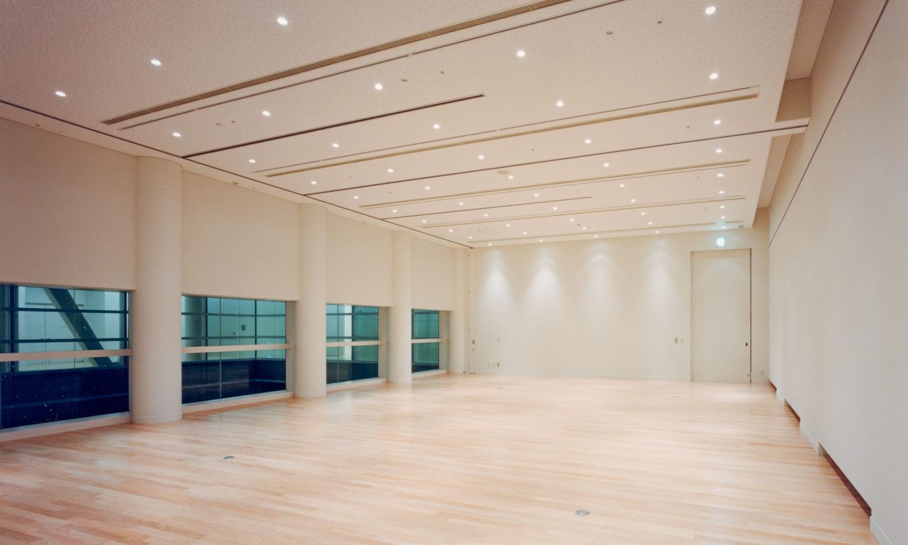 写真：白木の床に、白い壁の展示室。天井には移動壁のレールがある。ガレリア吹き抜けが見える窓がある。